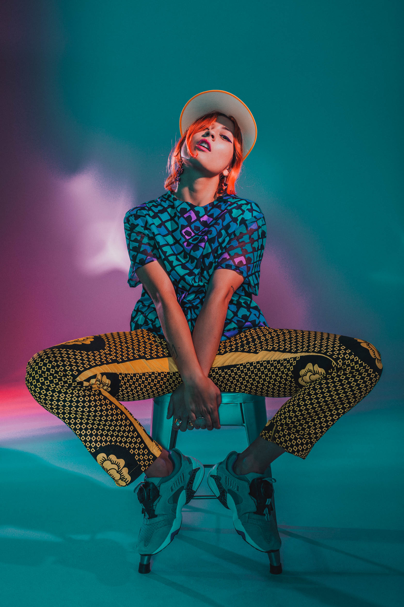 Stylish woman with orange hair poses on stool in a studio with teal and pink lighting, wearing a patterned top and pants.