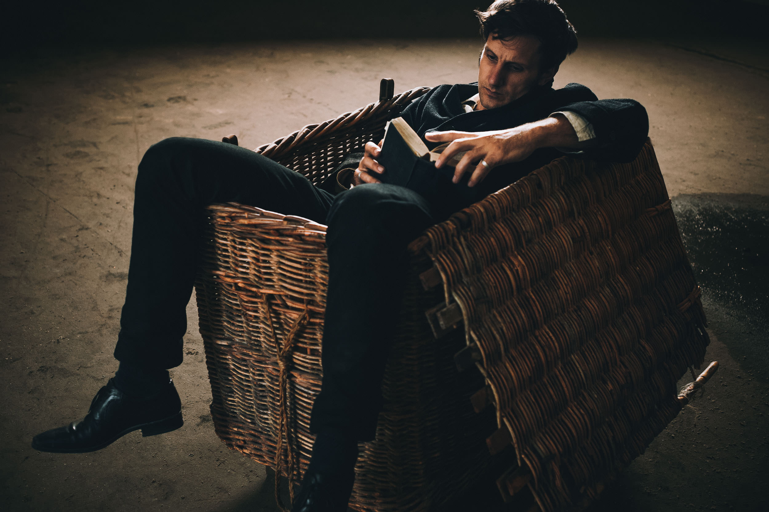 Man sitting in a wicker chair reading a book in a dimly lit room, wearing dark clothing and shoes, creating a contemplative atmosphere.
