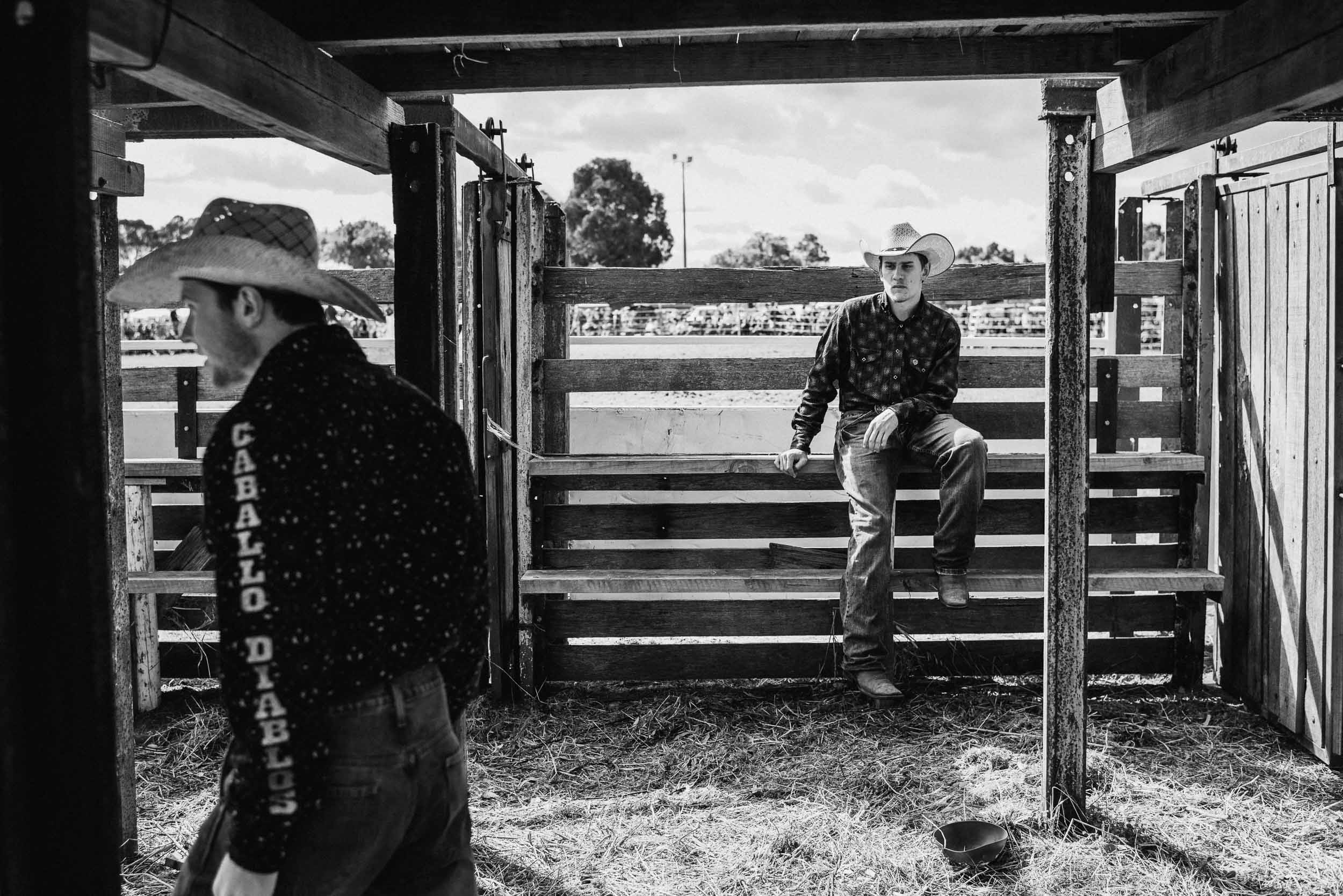 Two cowboys in black and white. One sits on a wooden fence, the other is walking toward the left side of the frame.