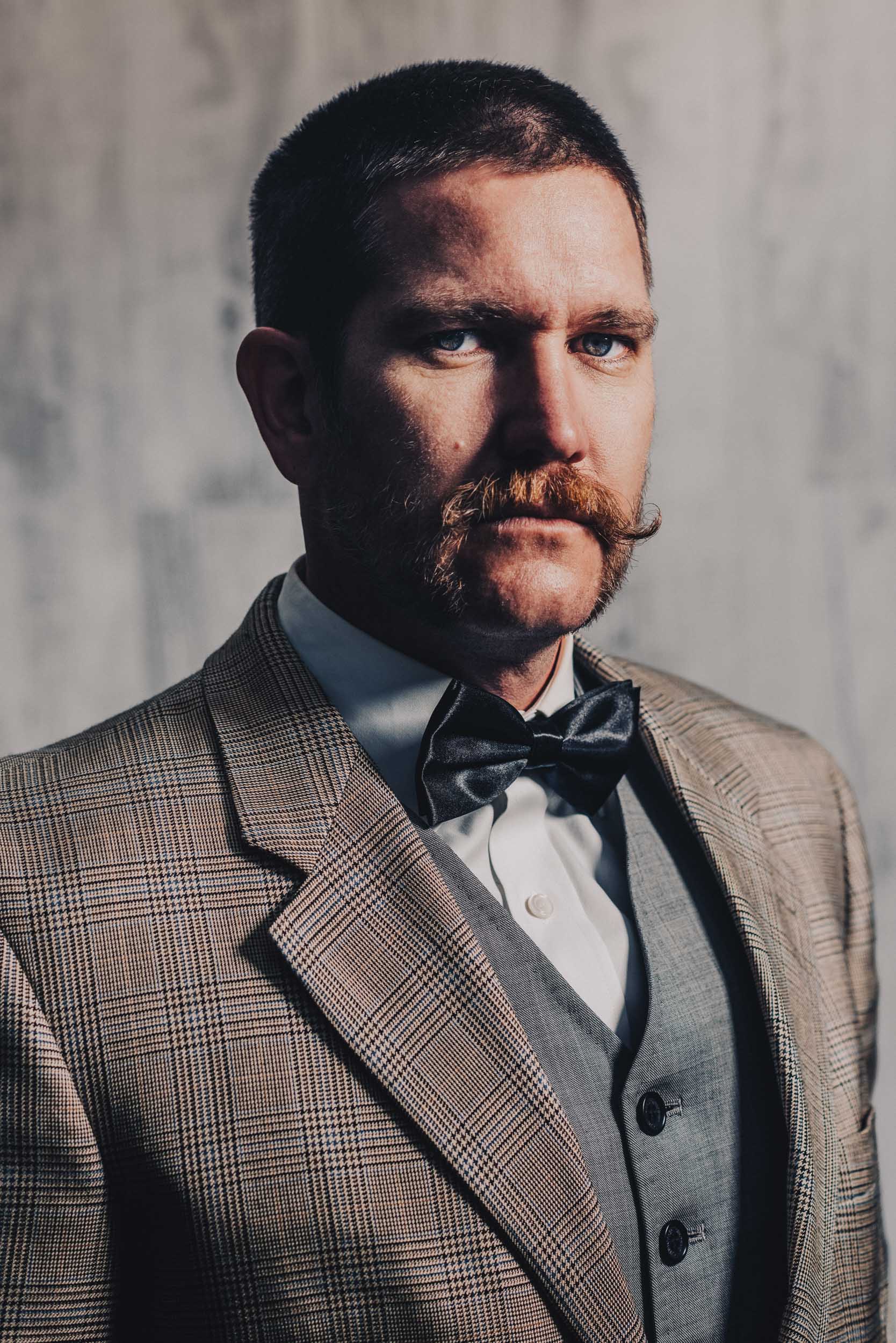 Portrait of an adult male with a handlebar mustache, wearing a suit with a bow tie, in front of a gray textured background.