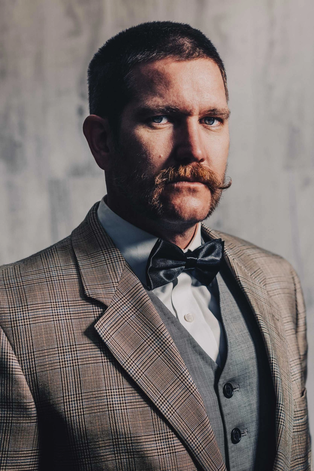 Portrait of a man with a mustache in a plaid jacket and bow tie against a textured wall by Wren Steiner.