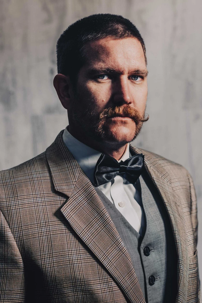 Portrait of a man with a mustache in a plaid jacket and bow tie against a textured wall by Wren Steiner.