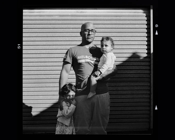Black and white portrait of a bald man wearing glasses holding a toddler while a young child stands beside him against a roller shutter.