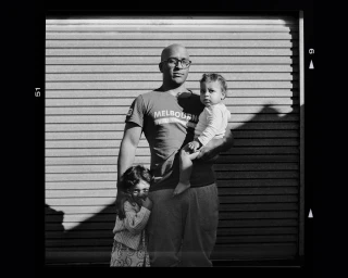 Black and white portrait of a bald man wearing glasses holding a toddler while a young child stands beside him against a roller shutter.