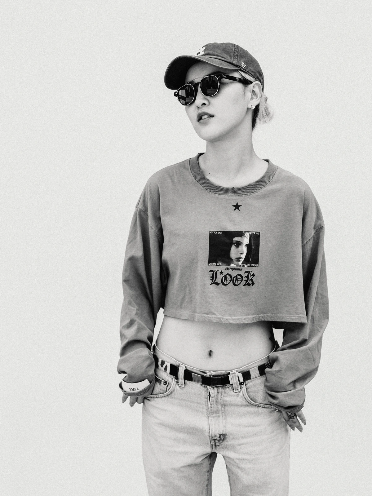 Monochrome portrait of a woman wearing a baseball cap, sunglasses, graphic crop top, and jeans against a plain background.