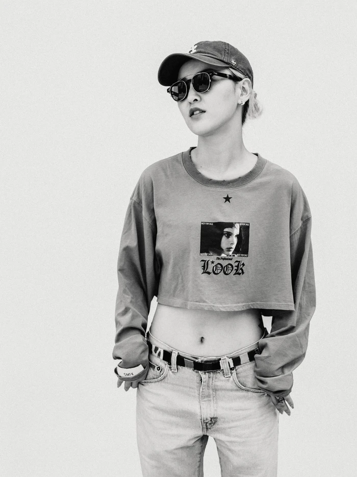 Monochrome portrait of a woman wearing a baseball cap, sunglasses, graphic crop top, and jeans against a plain background.