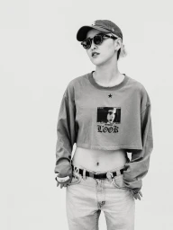 Monochrome portrait of a woman wearing a baseball cap, sunglasses, graphic crop top, and jeans against a plain background.