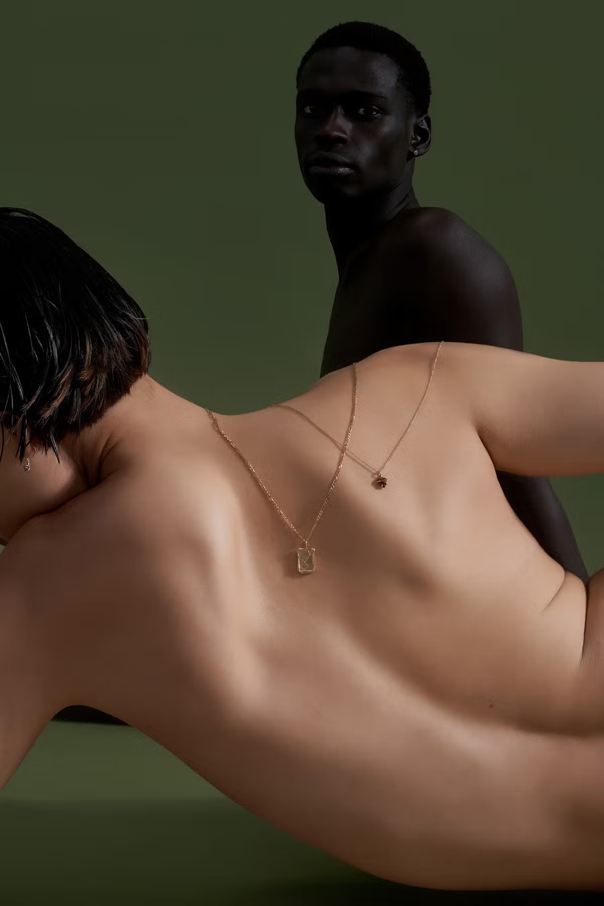 Two adults pose against a green backdrop, one lying down wearing delicate gold necklaces, the other standing and shirtless.