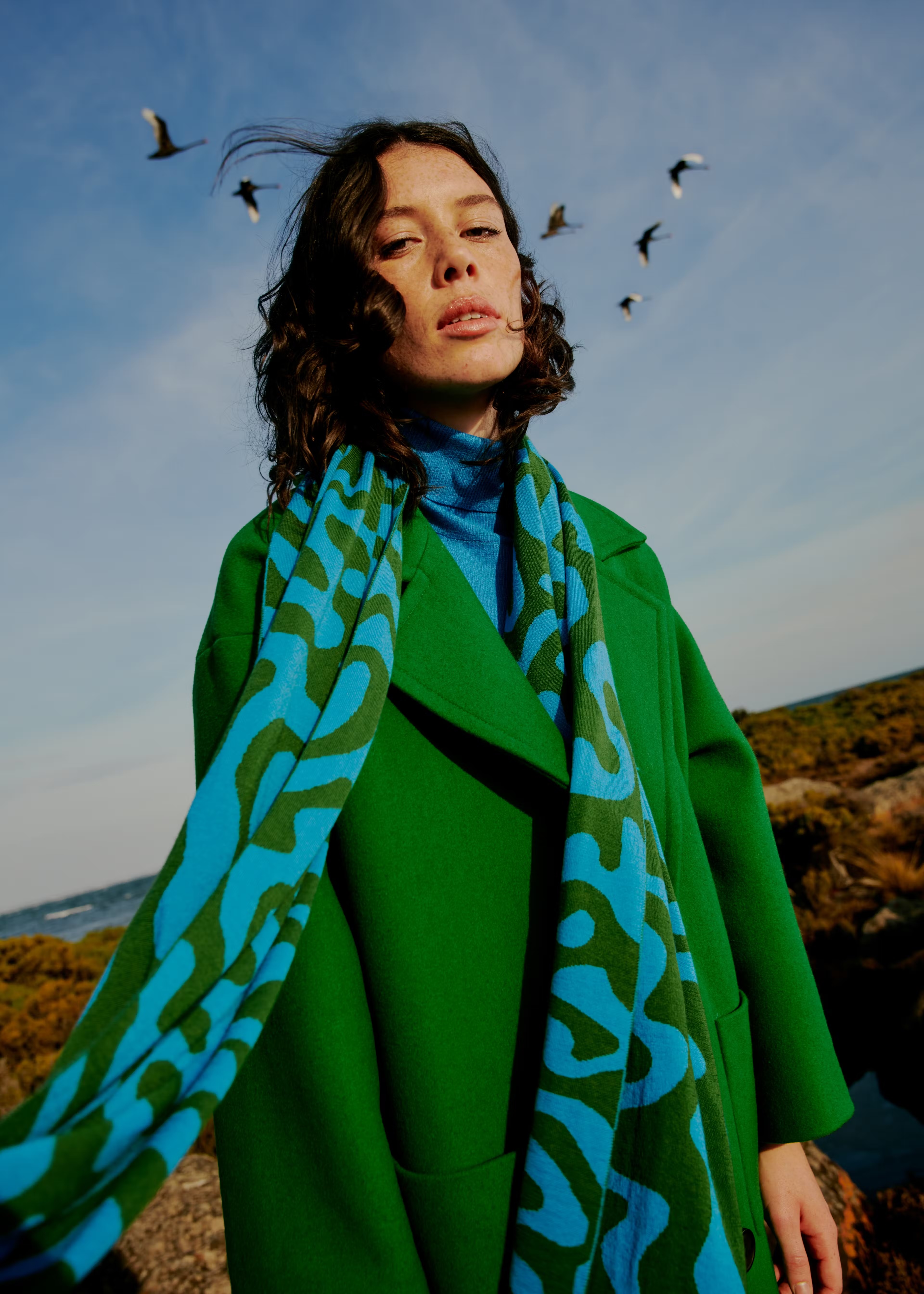 A young woman poses outdoors in a green coat and patterned scarf against a backdrop of birds and a blue sky with clouds.