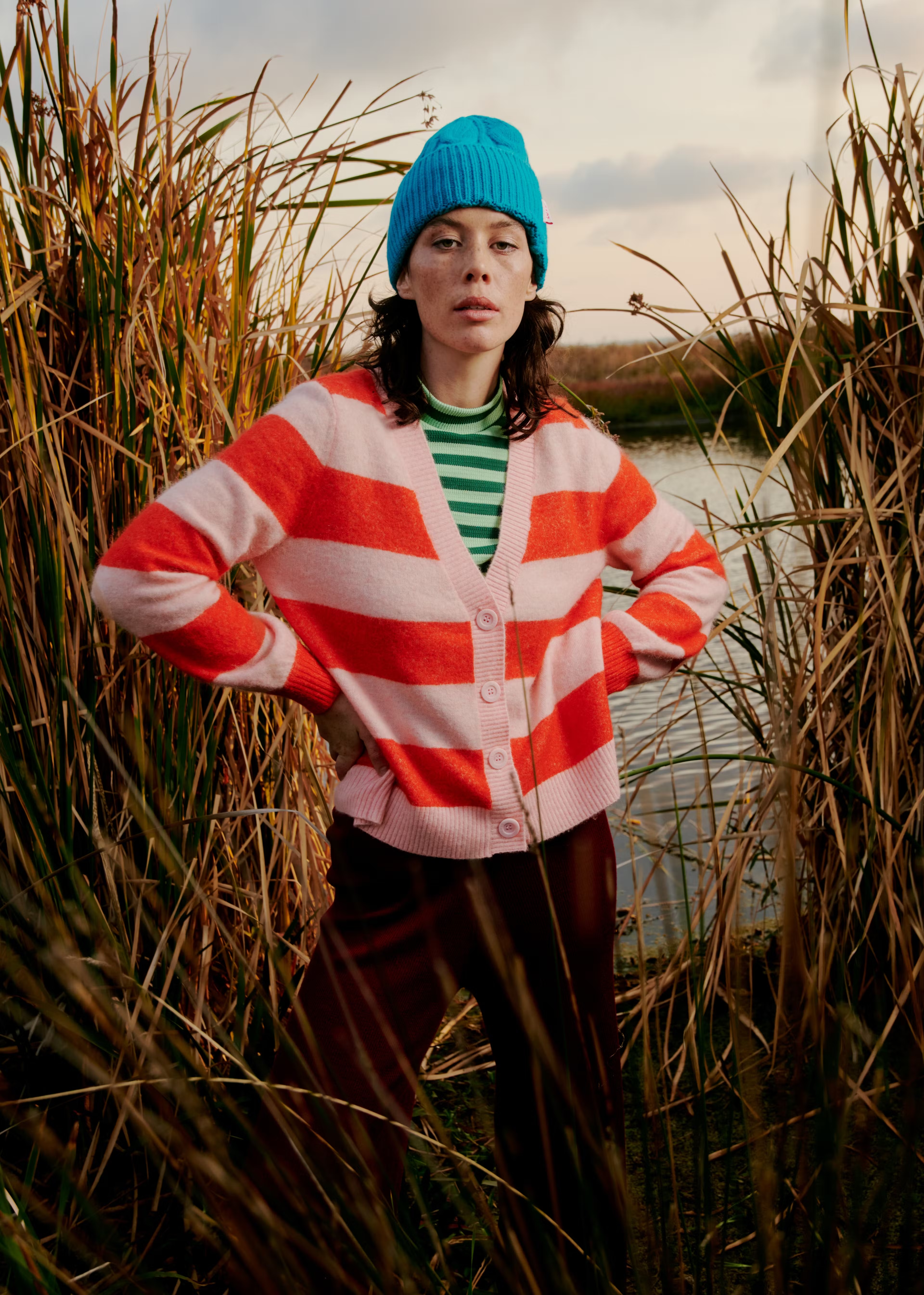 Adult woman in blue beanie and striped cardigan poses outdoors in tall reeds near a body of water during daytime.