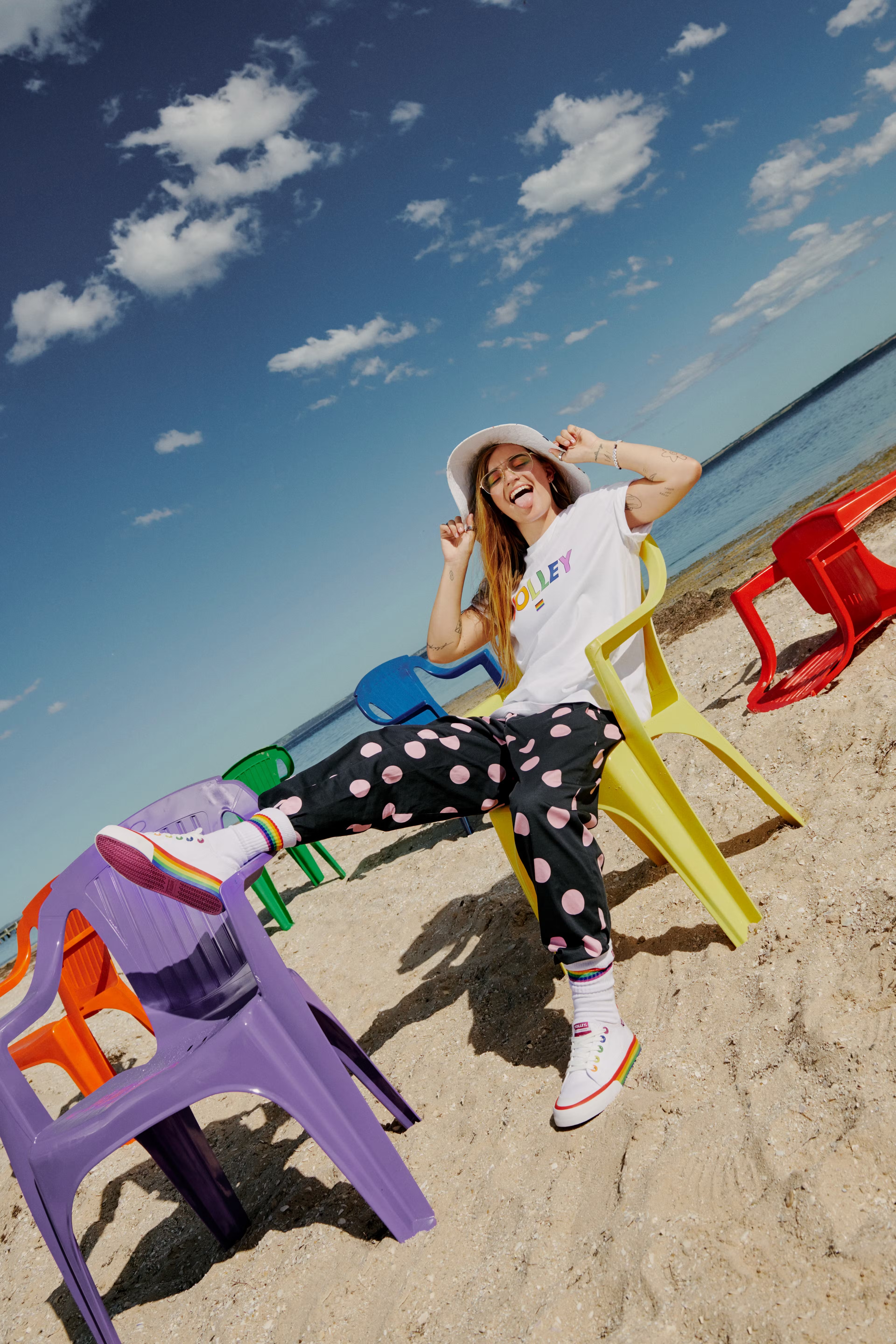 A young woman with long hair sits in a colorful plastic chair on a beach in Converse sneakers, black polka-dot pants, and a white T-shirt.