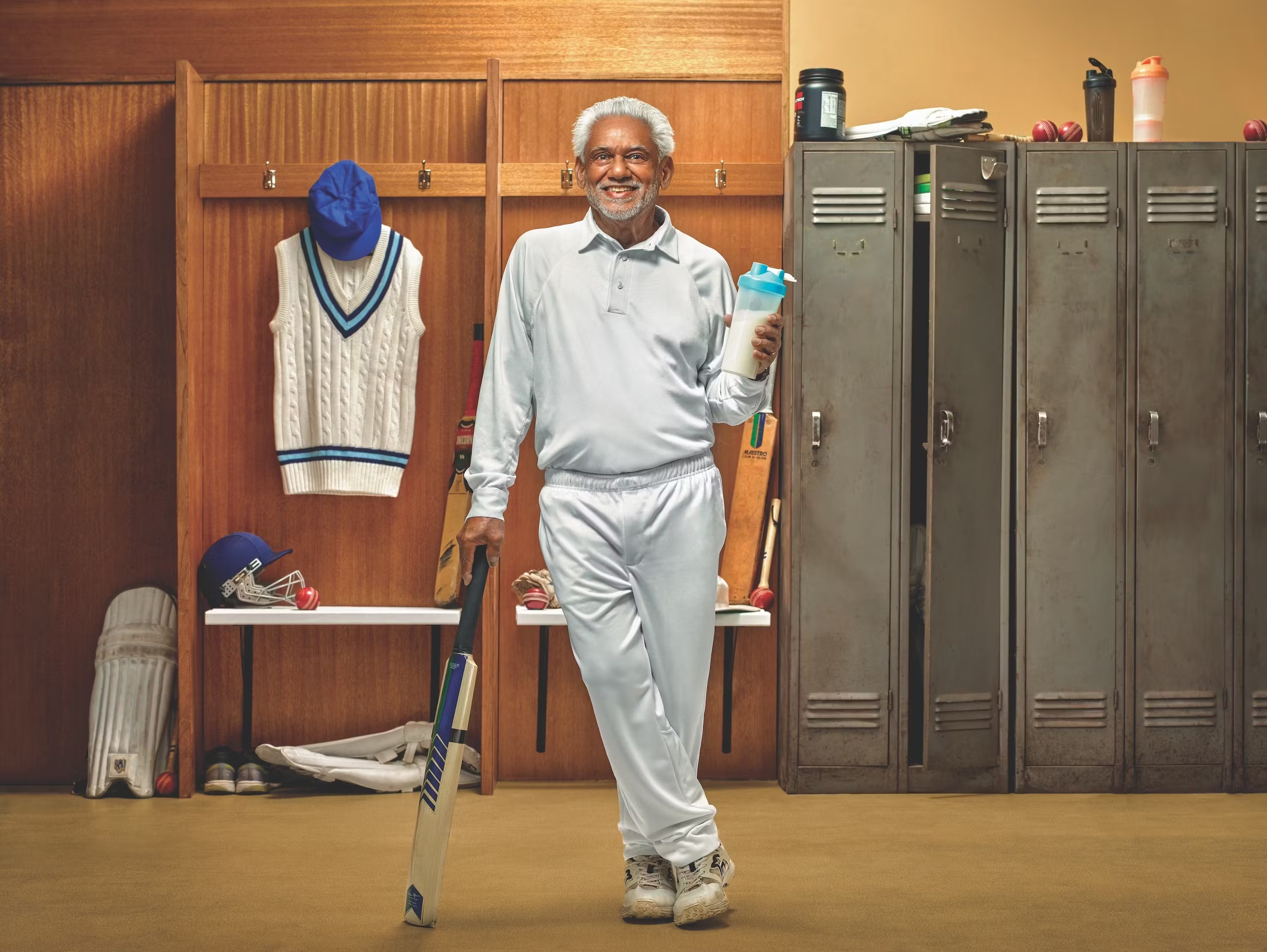 An elderly man smiles in cricket whites holding a protein shake and a cricket bat, standing in a locker room near sporting equipment.