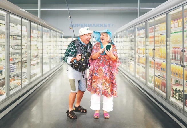 Elderly couple in a supermarket aisle, woman using a phone, man holding a fishing rod, creating a humorous, lifestyle scene.