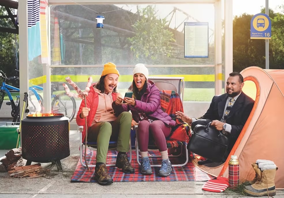 Two adults cheerfully look at a smartphone screen while seated next to a man in a business suit at an outdoor bus stop camping scene.