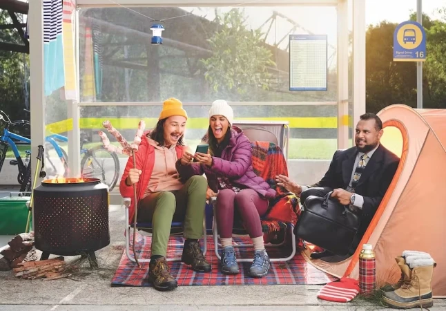 Two adults cheerfully look at a smartphone screen while seated next to a man in a business suit at an outdoor bus stop camping scene.