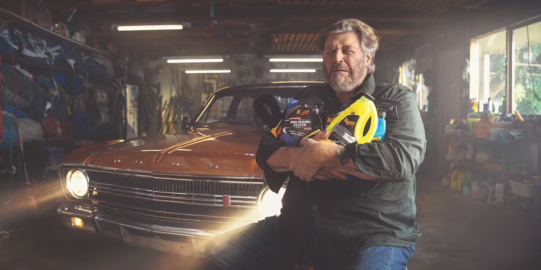 Man in garage smiles, holding Meguiar's car cleaning products in front of a vintage copper car, garage with overhead lights in background.