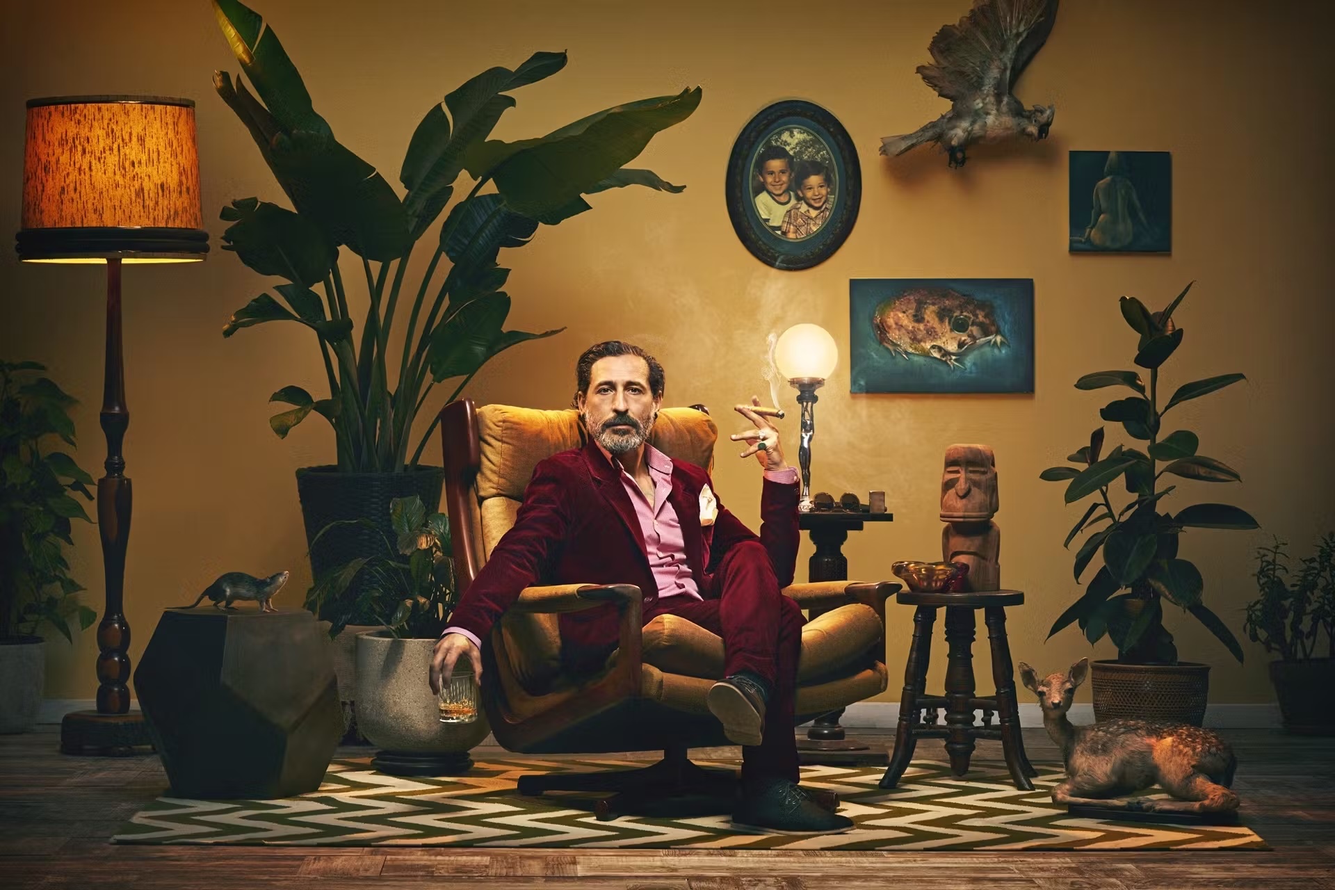 A man in a red suit sits in an armchair with a glass of whiskey and a cigar, surrounded by plants and retro decor in a warm, inviting room.