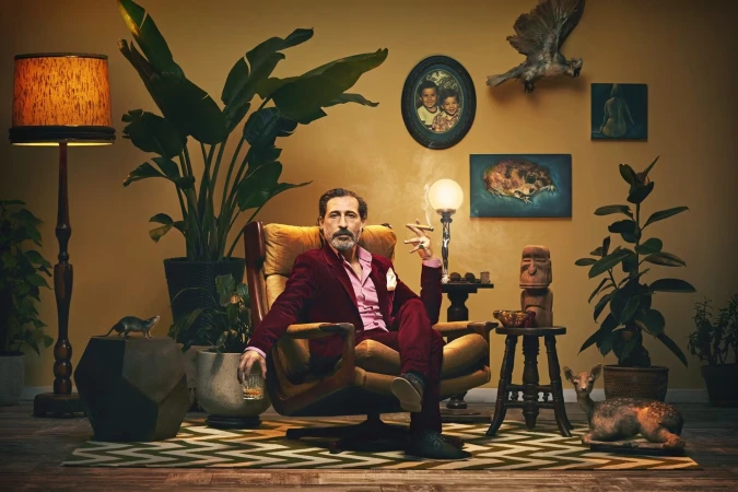 A man in a red suit sits in an armchair with a glass of whiskey and a cigar, surrounded by plants and retro decor in a warm, inviting room.