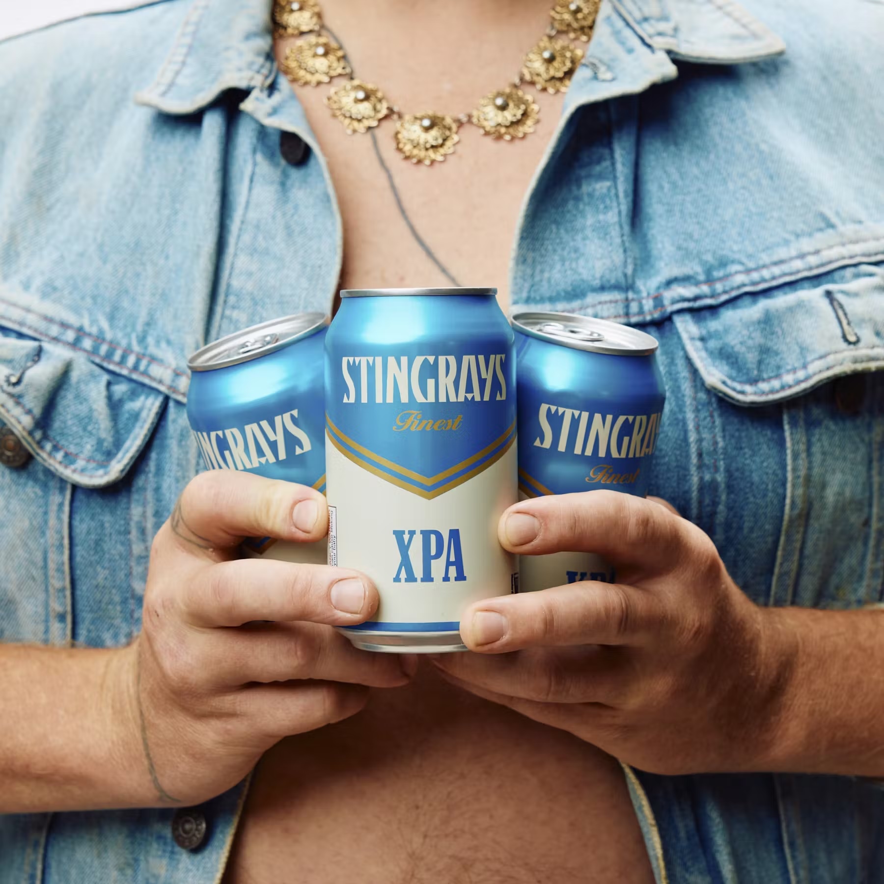 Three Stingrays Finest XPA beer cans are held by a person wearing a denim jacket and gold necklace, creating a refreshing and stylish image.