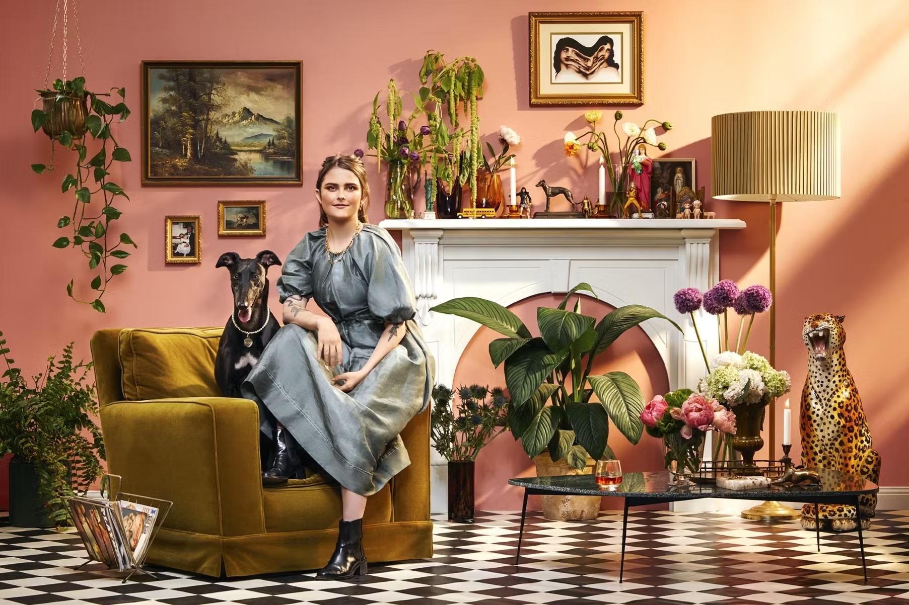 Woman in a blue dress poses with a greyhound on a mustard-colored armchair in an eclectic interior with art and plants.