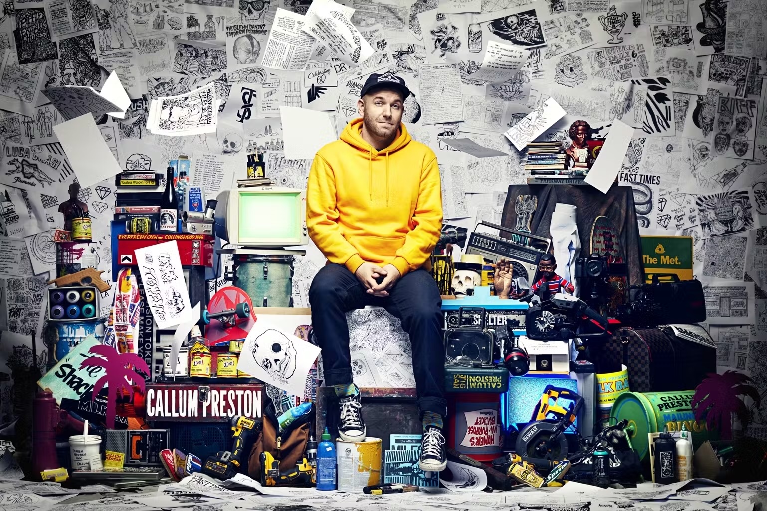 Young adult male in yellow hoodie seated amidst a collection of objects and artwork, surrounded by a backdrop of paper drawings.
