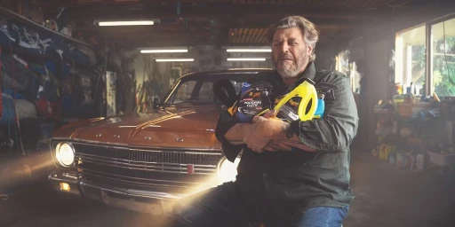 Middle-aged man holding car care products while leaning against a classic orange car in a garage