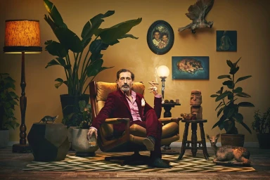 Middle-aged man in burgundy suit seated in armchair holding a cigar and glass, surrounded by eclectic vintage room decor.