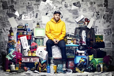 Man wearing a bright yellow hoodie and baseball cap seated amidst a cluttered array of art objects and sketches by Benny Capp