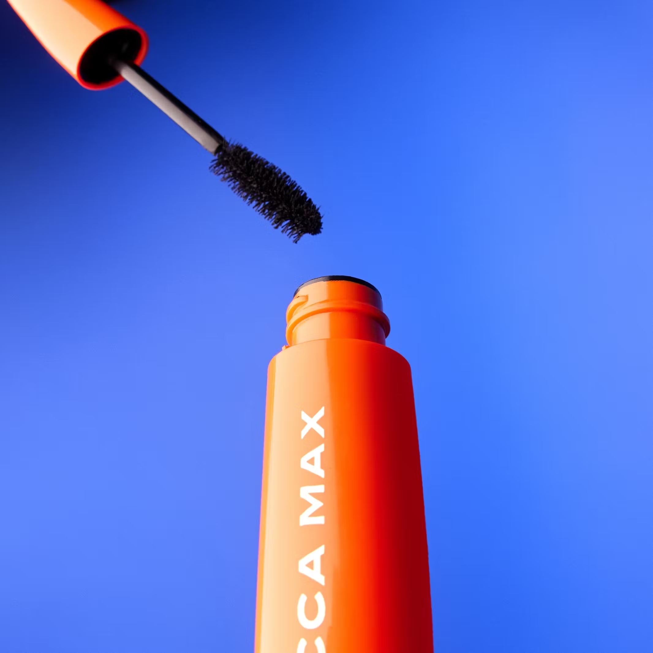 Close-up studio shot of orange tube of Becca Max mascara with black brush poised over the opening against a vibrant blue backdrop.