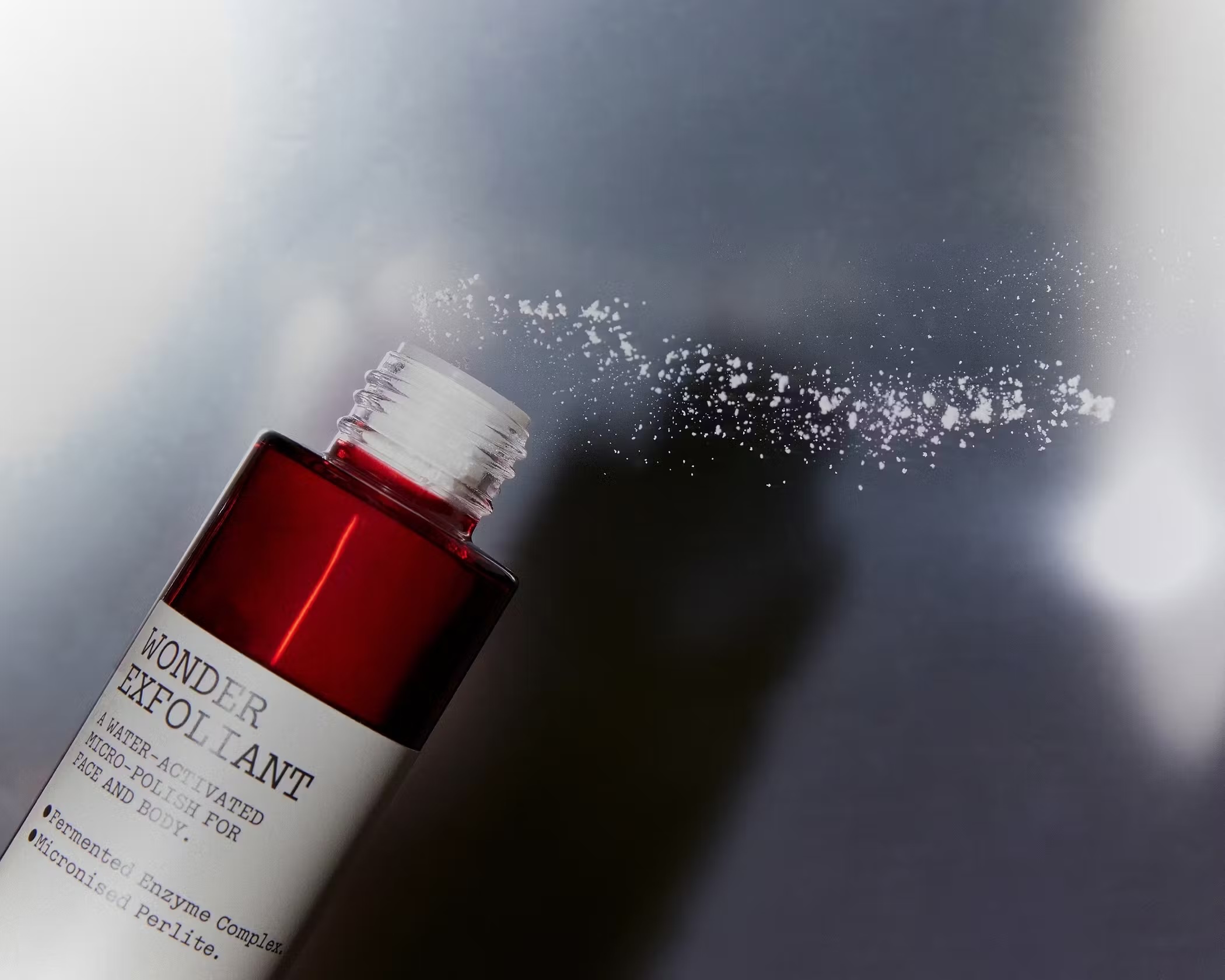 Close-up of an open bottle of red exfoliating polish with white specks of powder floating in the air against a muted background.