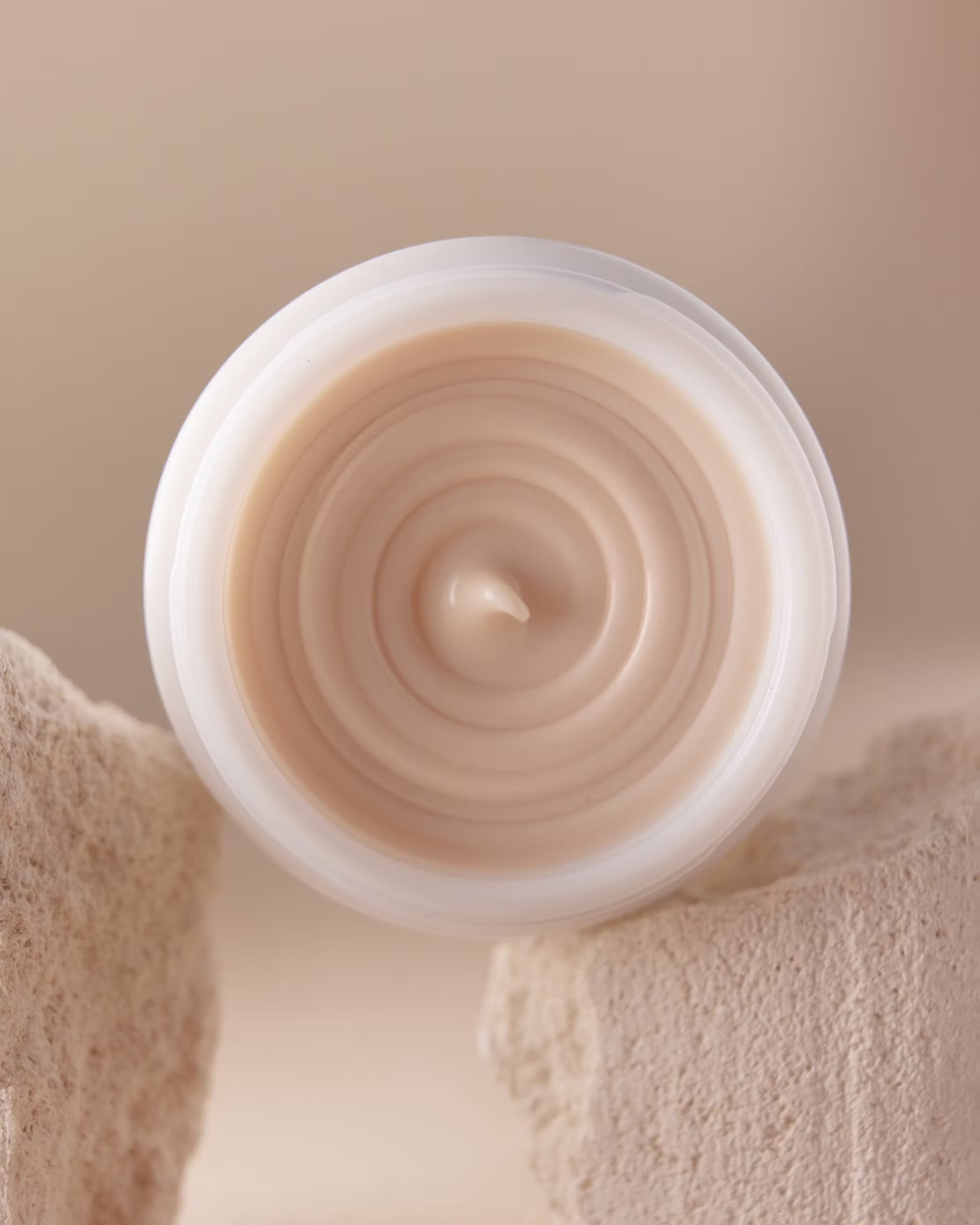 Top-down view of a jar of skin cream or lotion with white packaging and a textured stone block against a neutral background.