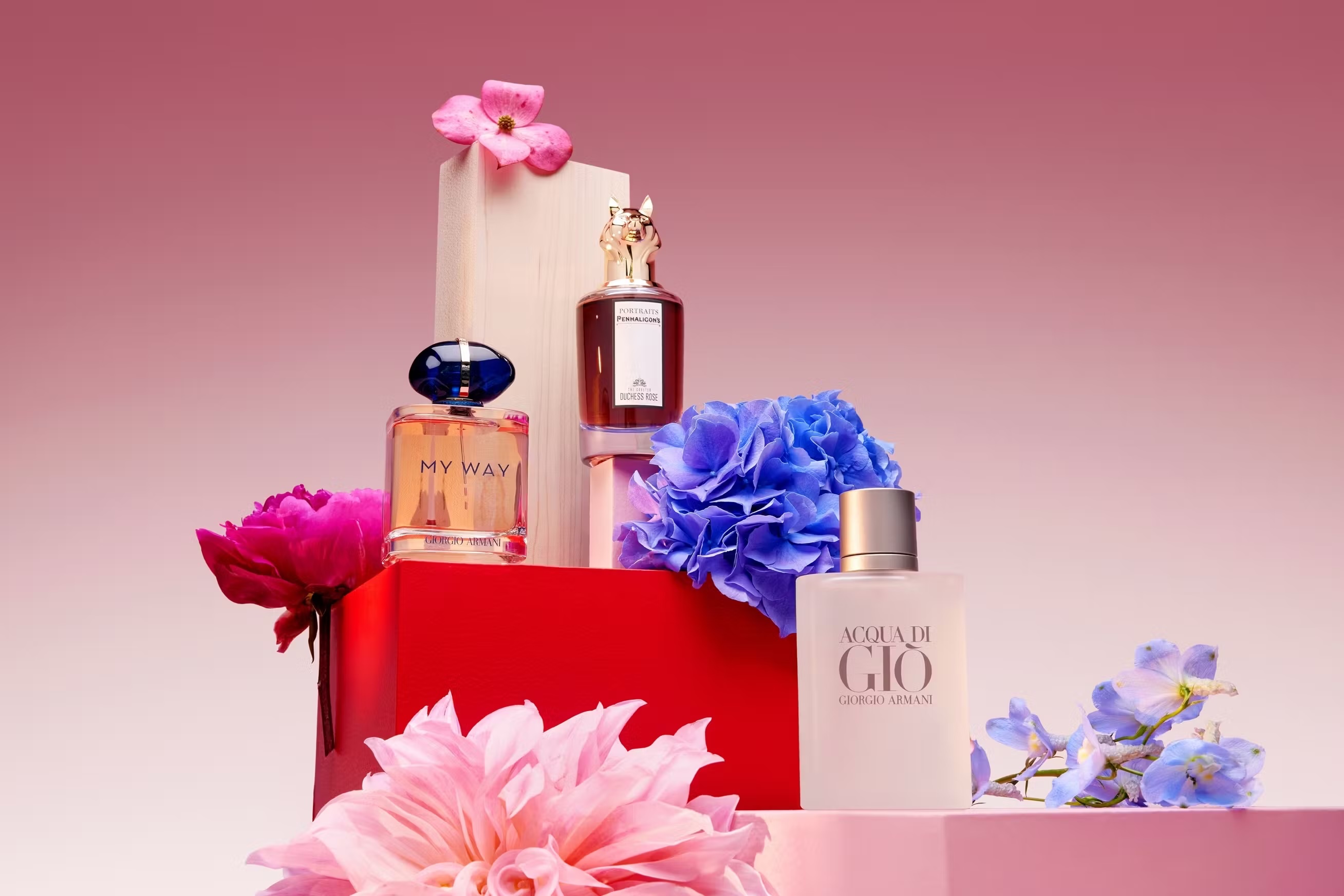Three perfume bottles are arranged on pink and red geometric blocks, surrounded by pink, blue, and purple flowers against a pink gradient backdrop.