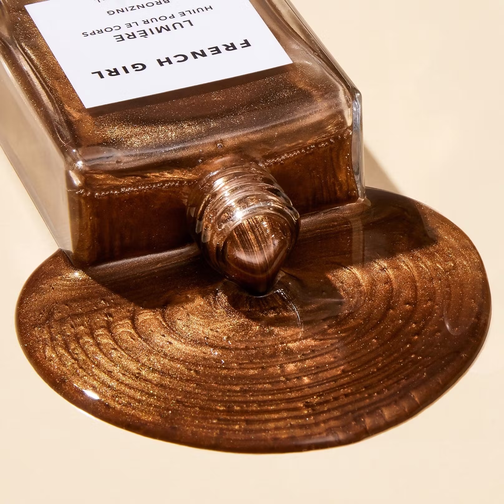 Close-up of a bottle of bronze colored French Girl Lumiere bronzing body oil with some of the product spilled onto a neutral surface.