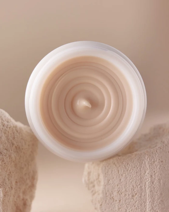 Top-down view of a round white jar containing swirled beige cosmetic cream nestled between textured neutral stone blocks.
