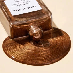 Close-up of a spilled glass bottle of shimmering bronze body oil pooled on a smooth beige surface.