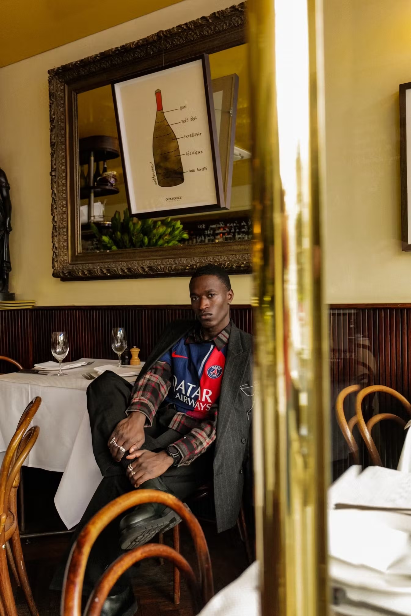 Man in a soccer jersey and blazer sitting at a restaurant table, with a wine bottle art print and mirror in the background.
