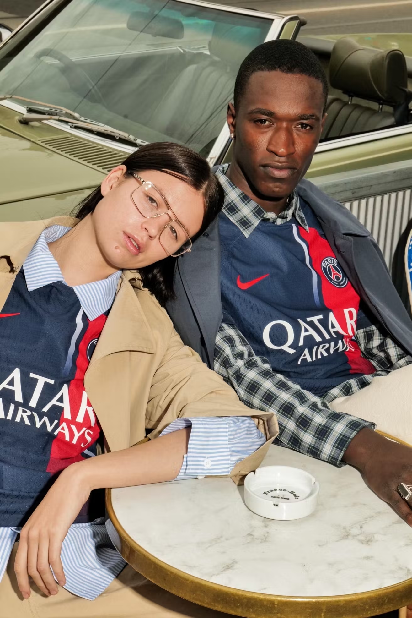 An Asian female and a Black male model sit in front of a light-green vintage convertible car wearing dark blue soccer jerseys with red and white accents.