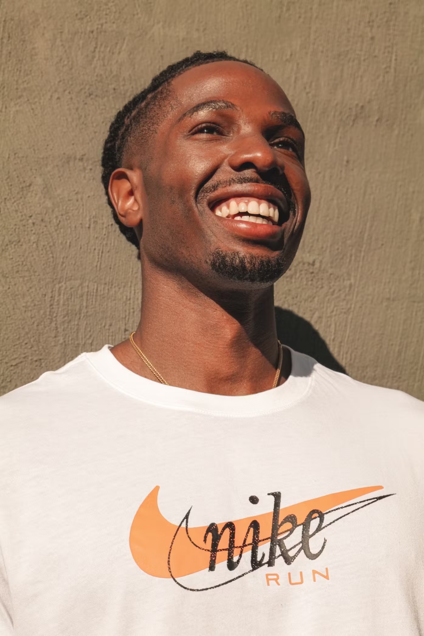 Portrait of a young Black man smiling and looking upward, wearing a white Nike T-shirt against a textured grey wall in soft sunlight.
