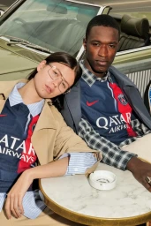 Two models wearing Paris Saint-Germain jerseys sitting outdoors at a round marble table near a vintage convertible car.