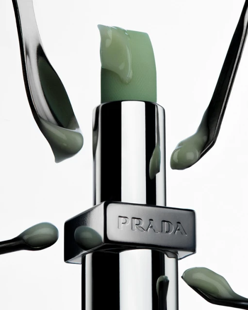 A tube of silver Prada lipstick stands vertically surrounded by spoons holding green balm, set against a clean white background.
