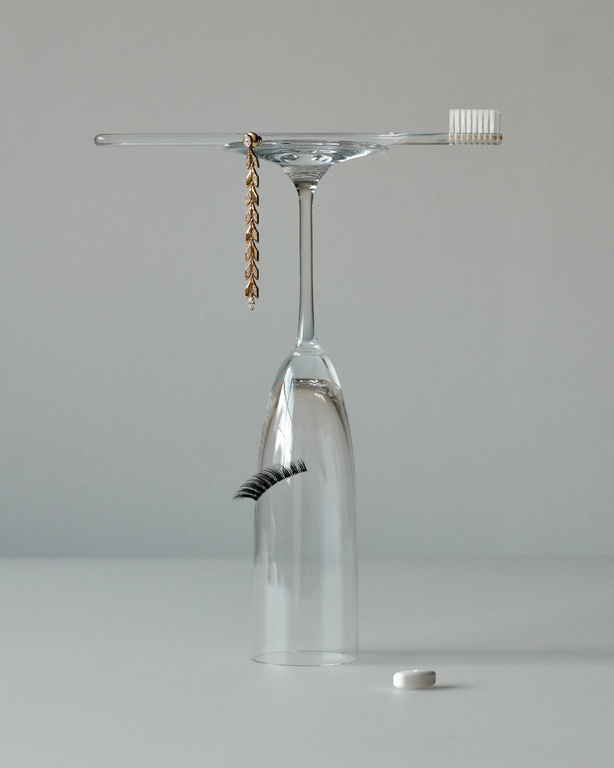 Still life shot of a toothbrush, gold chain, fake eyelash, and pill, all balanced atop a clear wine glass against a gray backdrop.
