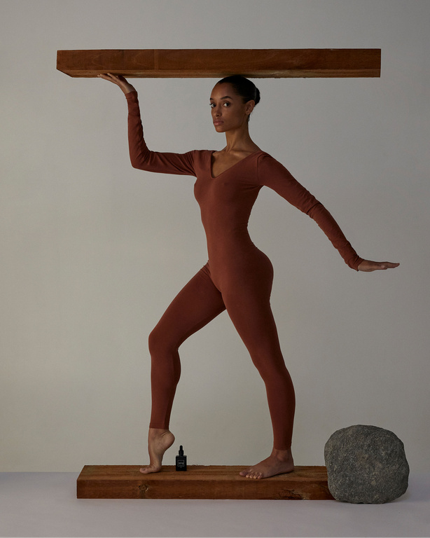 A slender woman in a brown jumpsuit balances a wooden plank above her head while standing on another. A bottle and rock are visible.