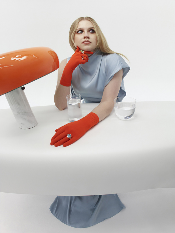 Blonde woman with red gloves seated at table with water glasses and retro orange lamp in a minimalist white setting, thoughtful pose.