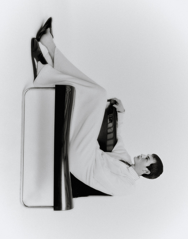 Monochrome fashion shot featuring an adult posing in white wide-leg pants and black heels on a chair against a white backdrop.