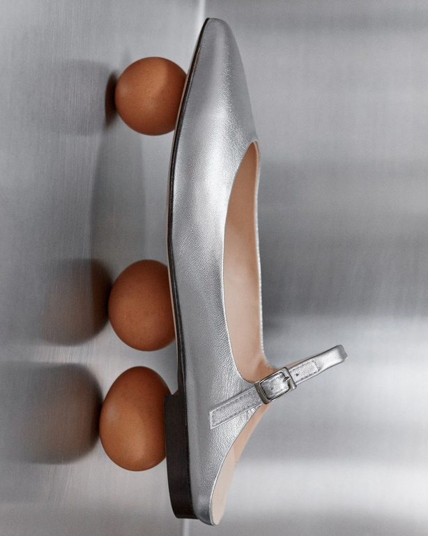 Elevated studio shot of a silver dress shoe with an ankle strap, accompanied by three brown chicken eggs against a silver backdrop.