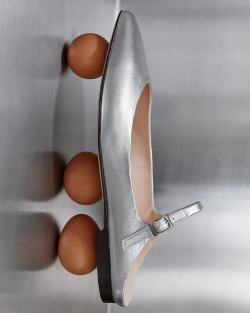 Elevated studio shot of a silver dress shoe with an ankle strap, accompanied by three brown chicken eggs against a silver backdrop.
