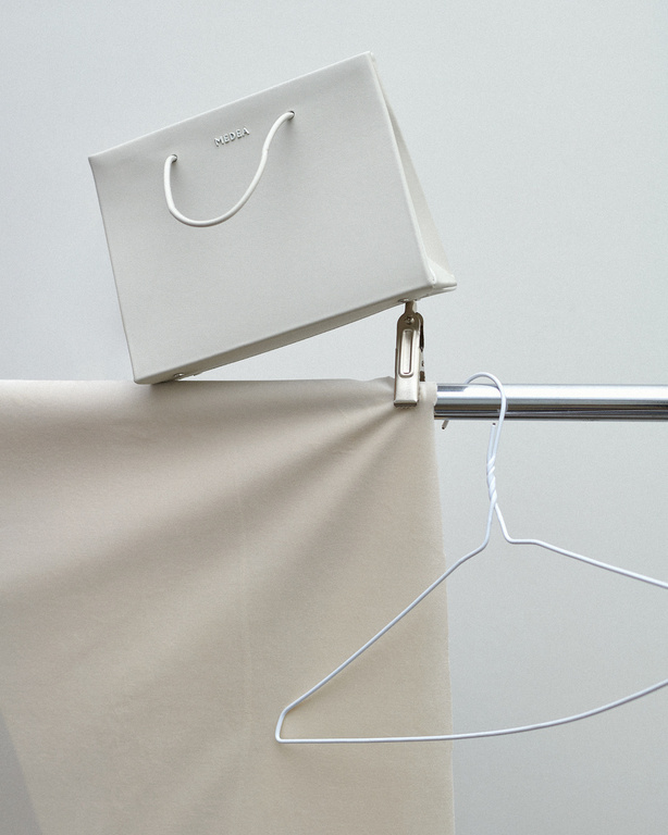Still life of a white paper shopping bag hanging from a metal clip on a clothing rack with a metal wire hanger, studio lighting.