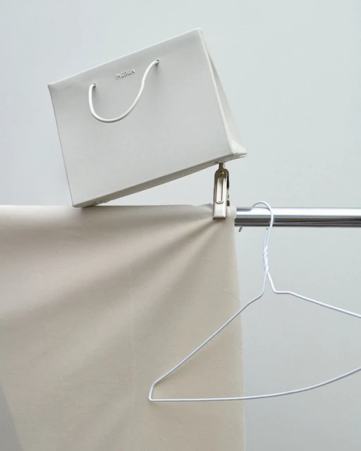 Still life of a white paper shopping bag hanging from a metal clip on a clothing rack with a metal wire hanger, studio lighting.