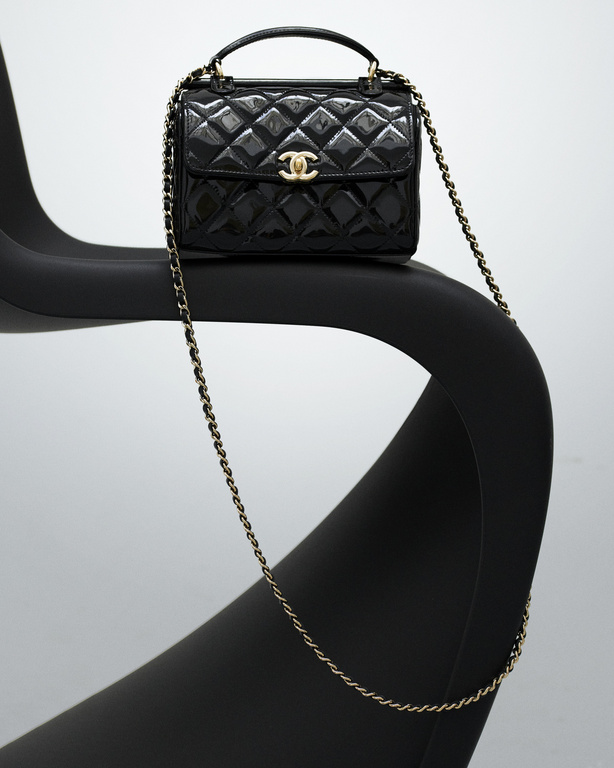 Black patent leather Chanel handbag with gold chain strap and handle on a black modern chair against a light grey background.