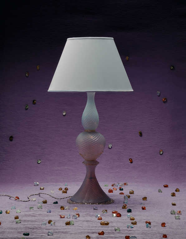 Illuminated glass table lamp with a white lampshade on a mauve textured background, surrounded by colorful decorative gems.